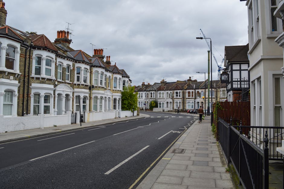 A quiet residential street in an urban area with a row of Victorian-style terraced houses featuring bay windows and brick facades, some with white-painted decorative elements. The street has a grey asphalt road with white lane markings, and a paved sidewalk with metal railings on the right side. An individual can be seen walking on the sidewalk in the distance. Overcast grey skies cast diffuse light over the scene. Visible are street lamps, a crane in the background, and parked cars along the curb. The scene depicts a typical neighbourhood environment suitable for home relocation, with no moving activities or furniture visible, but consistent with the setting where house removals, packing, and furniture transport might be performed by a company like Removals Elephant and Castle.