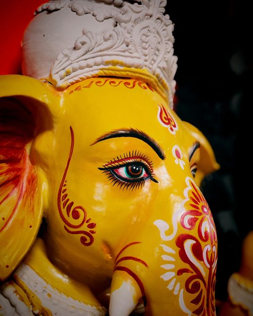 A close-up photograph of a traditional decorative statue of the Hindu deity Ganesha, featuring a bright yellow face with intricate red and white floral and geometric patterns. The statue has large, ex