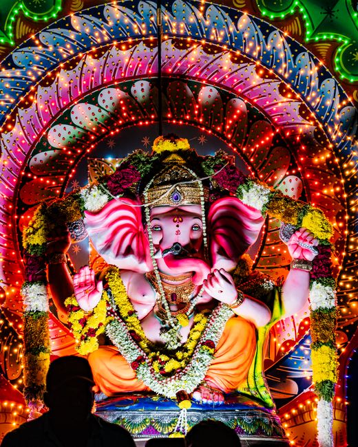 A close-up photograph of a traditional decorative statue of the Hindu deity Ganesha, featuring a bright yellow face with intricate red and white floral and geometric patterns. The statue has large, ex