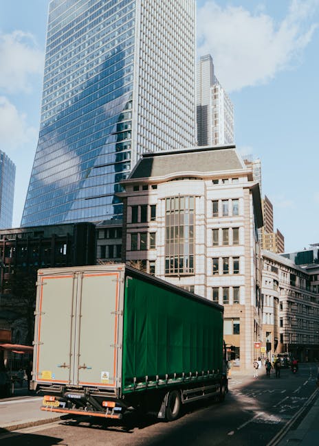 A large delivery truck with a green cargo container is driving along a city street in central London, passing tall modern glass office buildings and historic stone structures. The truck is in motion, with its rear doors closed and secured, and is situated near the curb with some pedestrians visible on the sidewalk. The scene depicts an urban environment with clear weather, blue sky, and sunlight illuminating the buildings, highlighting the busy logistics involved in house or office removals. The image reflects the transportation aspect of moving services offered by companies like Removals Elephant and Castle, fitting within their house and office relocation offerings.