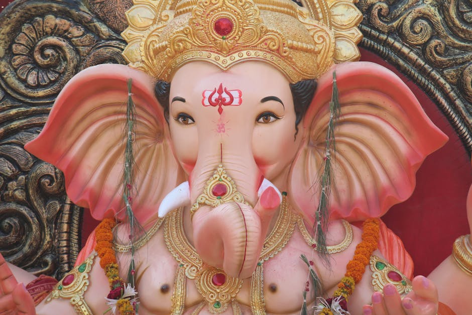 Close-up image of a decorated idol of Lord Ganesha, the Hindu elephant-headed deity, with a large pink and beige elephant head featuring big, flared ears. The idol is adorned with ornate gold jewelry, including a crown with red accents and various necklaces, and garlands of marigold flowers. The background showcases intricate carvings and decorative patterns in dark and gold tones. The idol is likely placed indoors, possibly in a temple or festive setting, surrounded by traditional decorations associated with religious or cultural celebrations. This detailed depiction emphasizes the craftsmanship and embellishments typical of Ganesha statues used during festivals, and the image highlights the importance of proper packing and handling in the context of moving or preserving such cultural artifacts, which can be overseen by professional removal services like Removals Elephant and Castle.