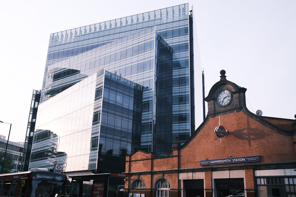 A large delivery truck with a green cargo container is driving along a city street in central London, passing tall modern glass office buildings and historic stone structures. The truck is in motion, with its rear doors closed and secured, and is situated near the curb with some pedestrians visible on the sidewalk. The scene depicts an urban environment with clear weather, blue sky, and sunlight illuminating the buildings, highlighting the busy logistics involved in house or office removals. The image reflects the transportation aspect of moving services offered by companies like Removals Elephant and Castle, fitting within their house and office relocation offerings.