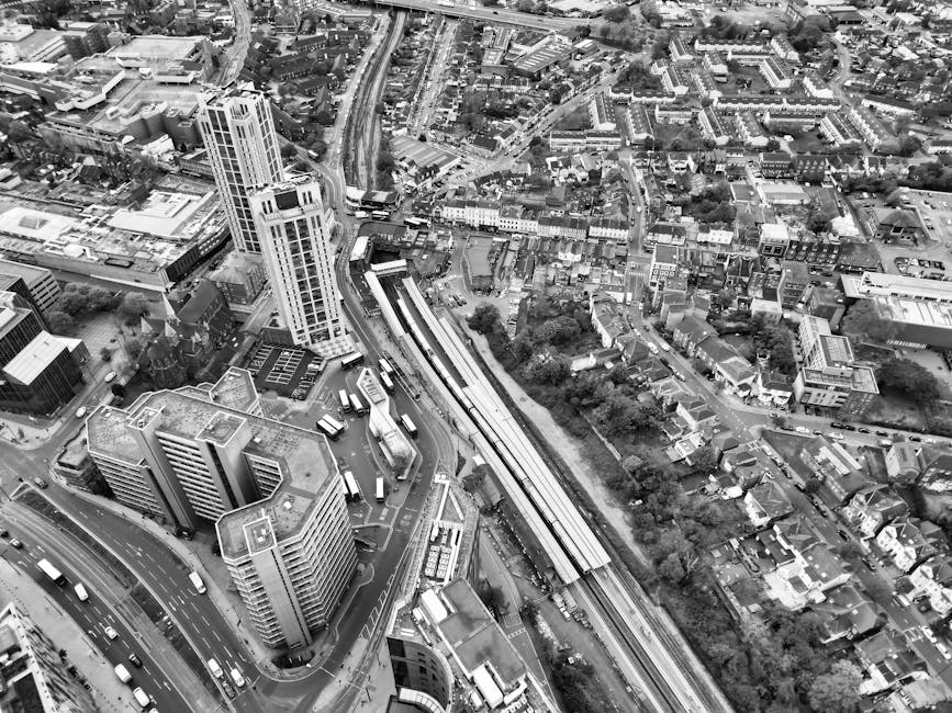 Black and white aerial photograph of the Elephant and Castle area showing multiple tall and mid-rise buildings clustered near the railway station, with train tracks, platforms, and train carriages visible alongside the surrounding urban landscape. The scene captures a busy city environment with roads, vehicles, and residential streets extending outward from the central station. The image illustrates an urban setting typical of London, with dense construction, commercial and residential structures, and greenery interspersed among the buildings. This view highlights the area's infrastructure, including the railway infrastructure integral to the home relocation and furniture transport services provided by Removals Elephant and Castle, as seen in the proximity of the station and the surrounding dense urban development.