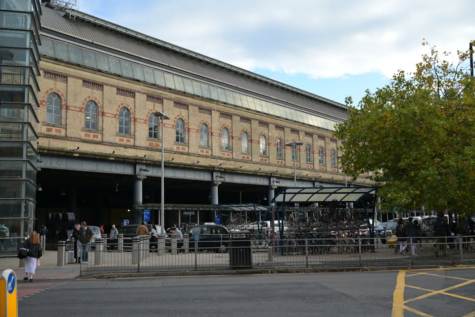 The image shows the exterior of a historic railway station building with ornate architectural details and a large arched entrance at ground level, located in the Elephant and Castle area. The building is constructed from light-colored stone and features decorative carvings, columns, and a parapet with the inscription 'Southern Railway' at the top. To the left of the entrance, there is a modern glass-fronted extension. Several people are visible outside, some walking and others standing near the entrance. The scene is illuminated by natural daylight under a clear blue sky. This setting illustrates the type of location where professional removals services, such as those provided by Removals Elephant and Castle, might operate during home relocation or furniture transport. The wide, paved forecourt in front of the building is used for easy access to load or unload items, highlighting the logistical considerations involved in packing and moving services.